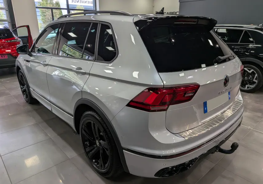 Vue 3/4 arrière droite d’un Volkswagen Tiguan 1.5 TSI R-Line 2024 en reflet d’argent avec jantes noires et attelage.