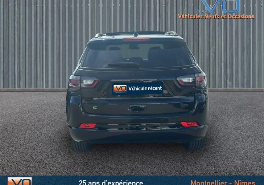 Vue arrière d'un Jeep Compass noir 2023 avec barres de toit et feux arrière LED allumés sur fond industriel.