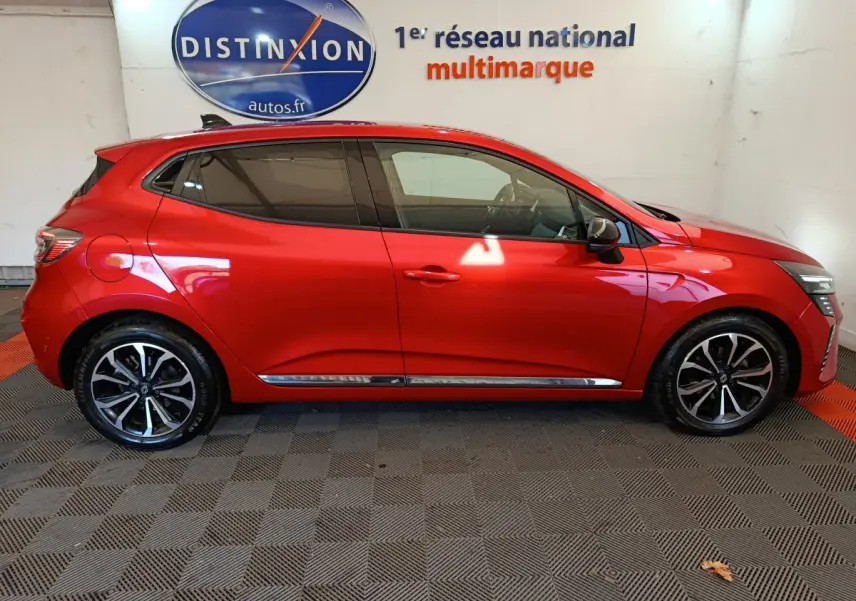 Profil droit d'une Renault Clio Techno TCe 90 rouge 2024 avec jantes alliage et vitres teintées en intérieur showroom.