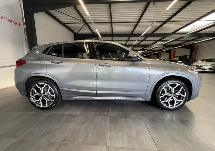BMW X2 sDrive 18d gris foncé vue de profil côté gauche avec jantes alliage 19 pouces style 722 M en intérieur showroom.