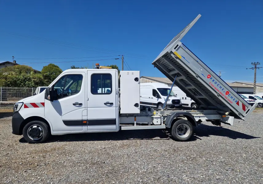 Profil côté gauche d’un Renault Master blanc avec benne relevée et coffre latéral visible sur un sol gravillonné.
