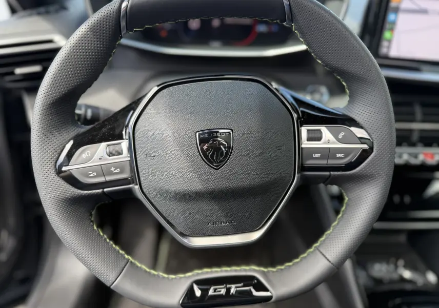 Gros plan sur le volant cuir perforé noir avec surpiqûres vertes du Peugeot 2008 Hybrid 145ch GT, tableau de bord en arrière-plan flou.
