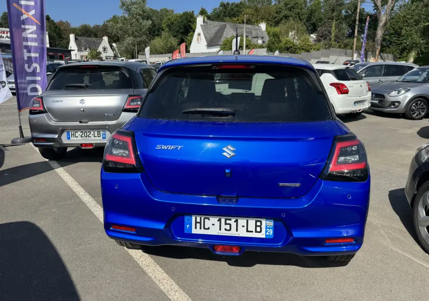 Vue arrière d'une Suzuki Swift 1.2 Privilège Hybrid bleue, stationnée en extérieur sur un parking.