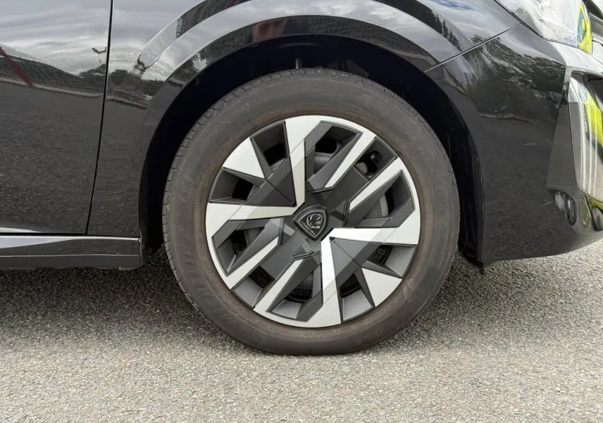Gros plan sur la roue avant droite d’une Peugeot 208 noire avec jante au design géométrique bicolore.