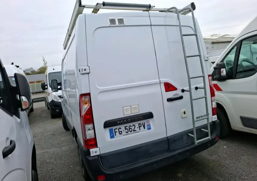Vue arrière d’un Renault Master blanc avec galerie et échelle métallique, stationné entre deux autres véhicules utilitaires.
