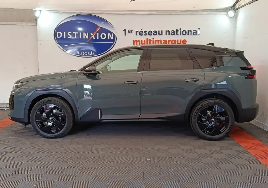 Vue de profil côté gauche d'une Citroën C5 hybride 2026 vert foncé avec jantes noires brillantes en intérieur showroom.
