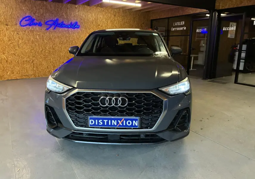 Audi Q3 Sportback gris vue de face avec calandre hexagonale noire et phares LED allumés en intérieur showroom.