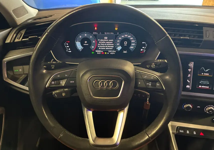 Vue intérieure centrée sur le volant cuir multifonctions de l'Audi Q3 Sportback gris avec tableau de bord numérique allumé.