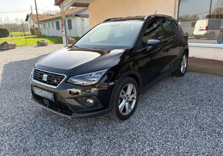 SEAT Arona noir vue 3/4 avant droit, avec calandre hexagonale et jantes alliage, stationné sur gravier devant un bâtiment.