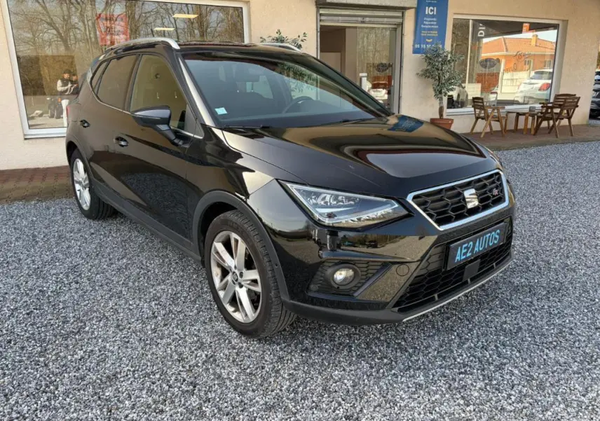 SEAT Arona noir vue 3/4 avant droit, avec calandre hexagonale et jantes alliage, stationné sur gravier devant un bâtiment.