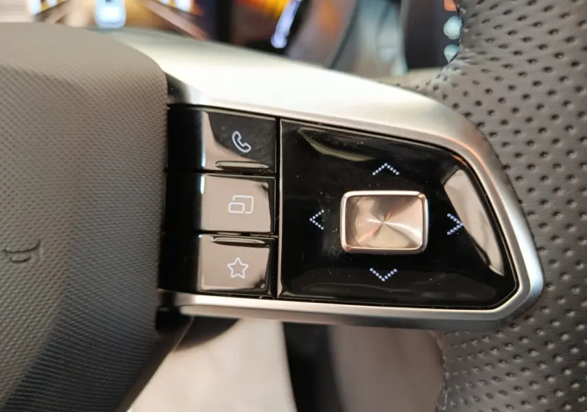 Gros plan sur les commandes gauche du volant perforé noir du MG ZS blanc, avec boutons multifonctions et finition argentée.