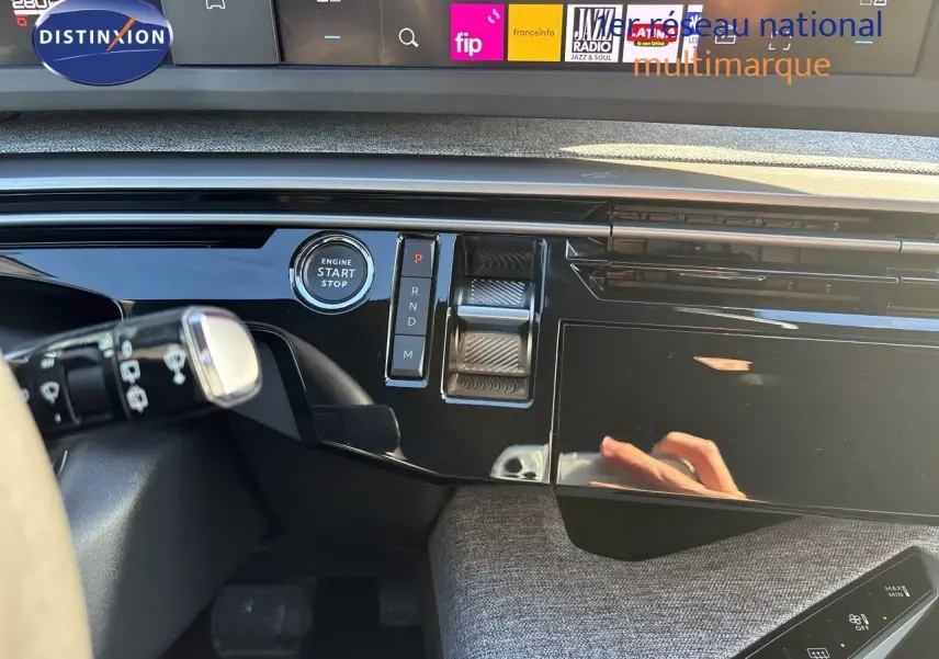 Vue rapprochée de la console centrale noire brillante du Peugeot 5008 hybride 2025, avec bouton start/stop et commandes de boîte auto.
