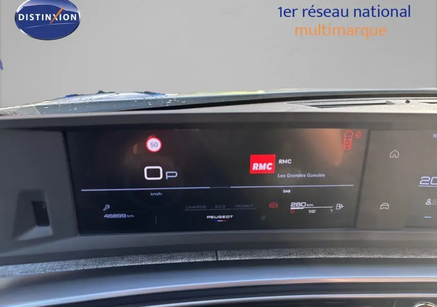 Tableau de bord numérique du Peugeot 5008 blanc, affichant la vitesse, la radio RMC et l'autonomie.