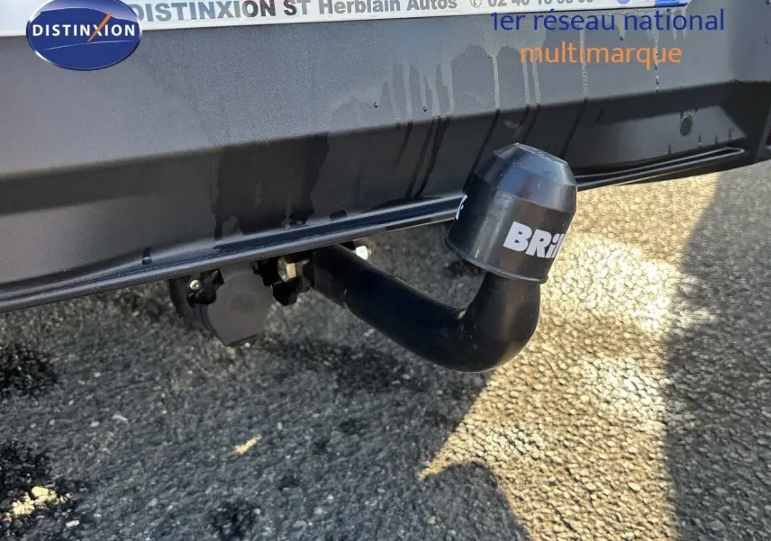 Gros plan sur l'attelage noir fixé à l'arrière d'un Peugeot 5008 blanc Okenite, avec fond de sol goudronné.