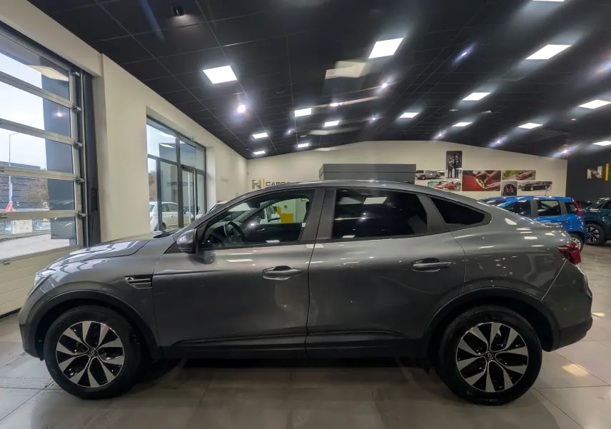 Vue latérale droite d'un Renault Arkana gris métal 2022 en hybride essence, avec jantes bi-ton et vitres teintées, en showroom.