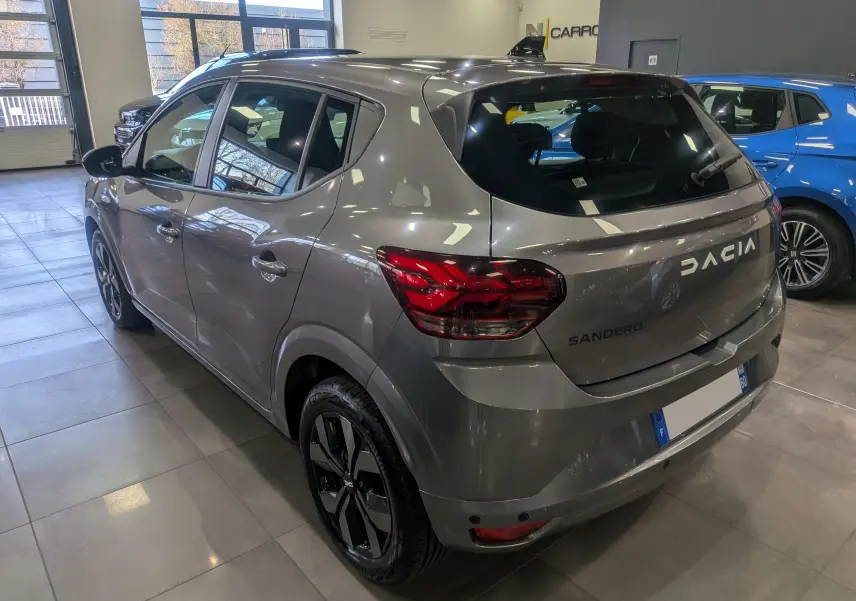 Vue 3/4 arrière droite d'une Dacia Sandero TCe 90 gris schiste en showroom, avec jantes noires distinctives.