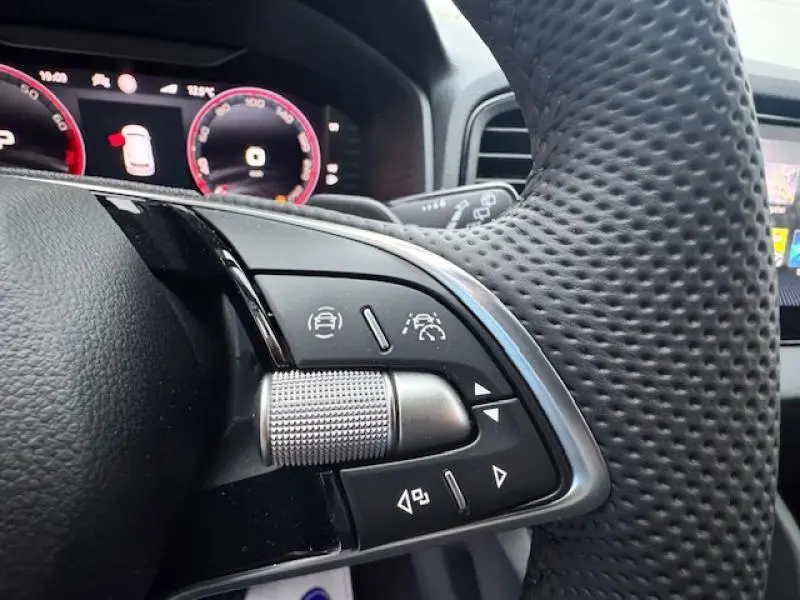 Gros plan sur les commandes du volant à droite en cuir perforé du Skoda Karoq gris, avec tableau de bord numérique en arrière-plan.