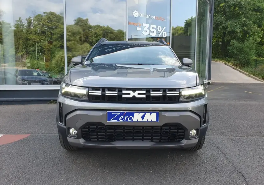 Vue avant d'un Dacia Duster 2026 gris schiste avec calandre noire et phares allumés, devant une vitrine de concession.