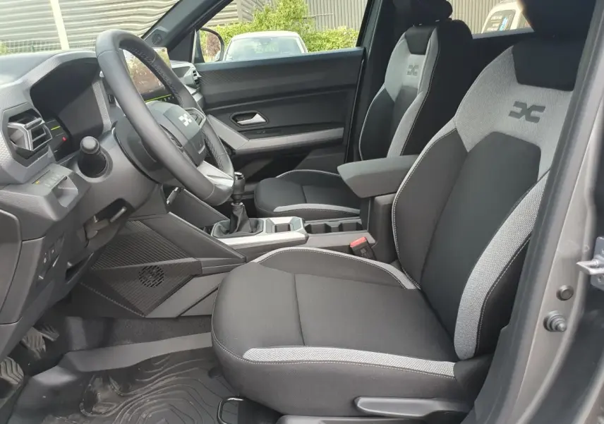 Vue intérieure côté conducteur du Dacia Duster 2026, sièges tissu noir et gris avec logo distinctif brodé.