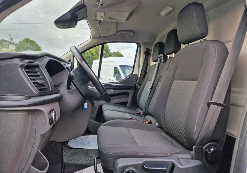 Vue intérieure côté conducteur du Ford Transit Custom blanc, montrant les sièges avant en tissu gris et le volant noir.
