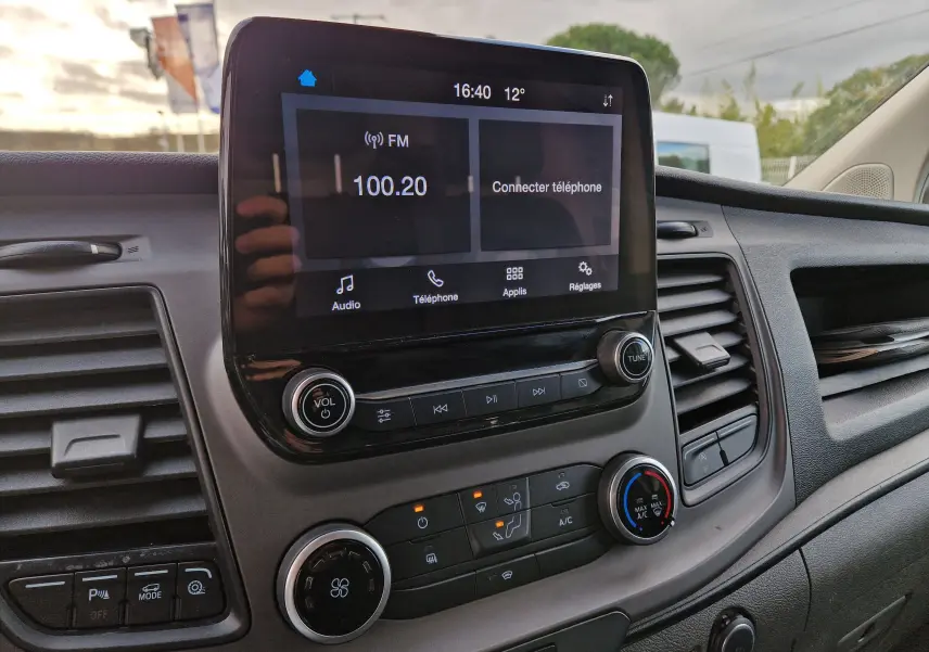 Vue intérieure du tableau de bord du Ford Transit Custom blanc, avec écran tactile central et commandes climatisation visibles.