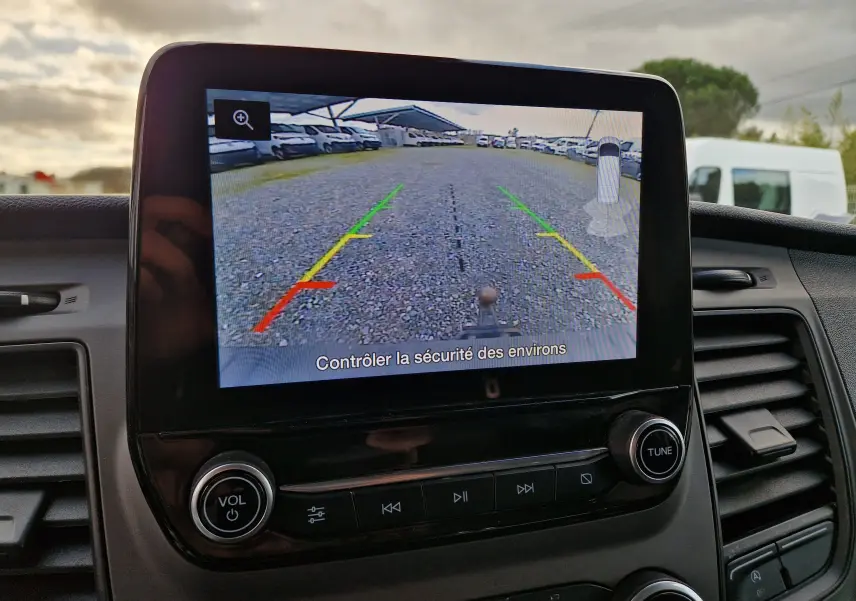 Écran central du Ford Transit Custom blanc montrant la caméra de recul avec lignes de guidage colorées sur un sol gravillonné.