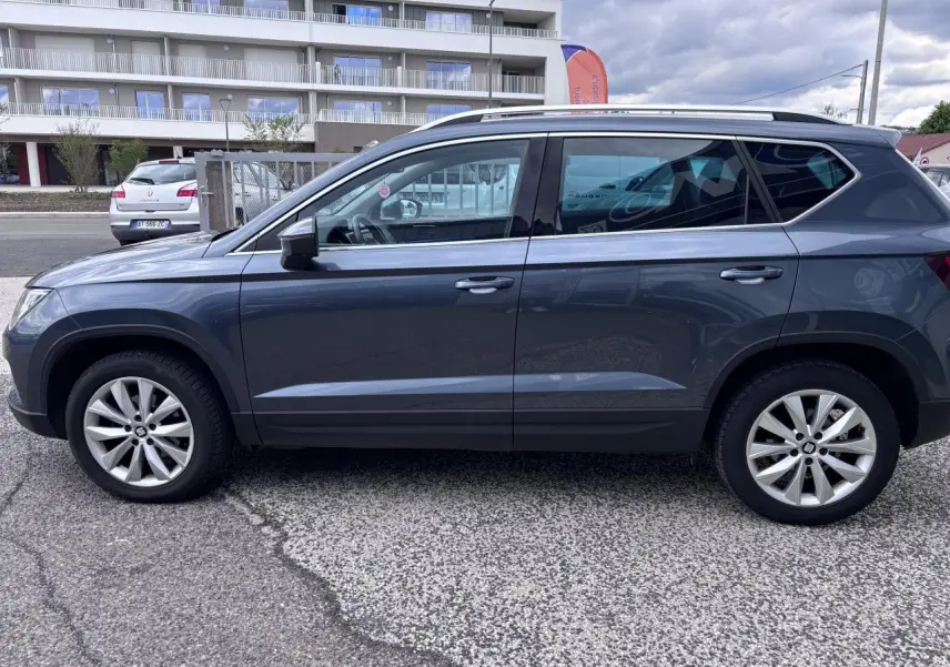 Vue latérale droite d'un SEAT Ateca gris foncé 2019 avec jantes alliage 17'' et barres de toit chromées.