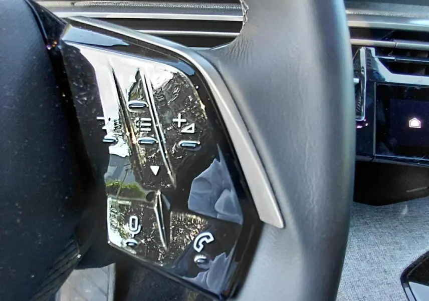 Gros plan sur les commandes au volant côté gauche du Peugeot 5008 gris Hybrid 2025, avec écran tactile en arrière-plan.