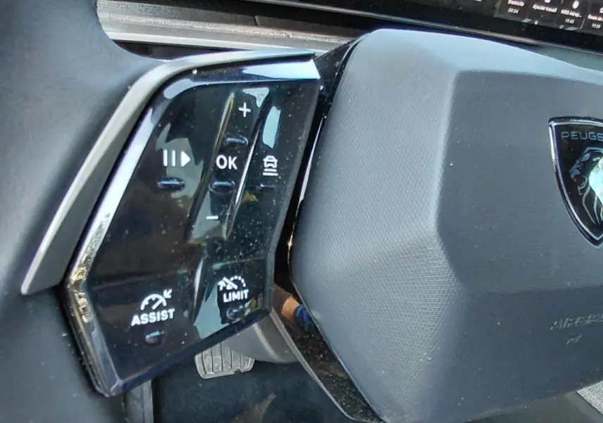 Gros plan sur le volant gris du Peugeot 5008 Hybrid 2025, avec commandes multifonctions et logo central visible.