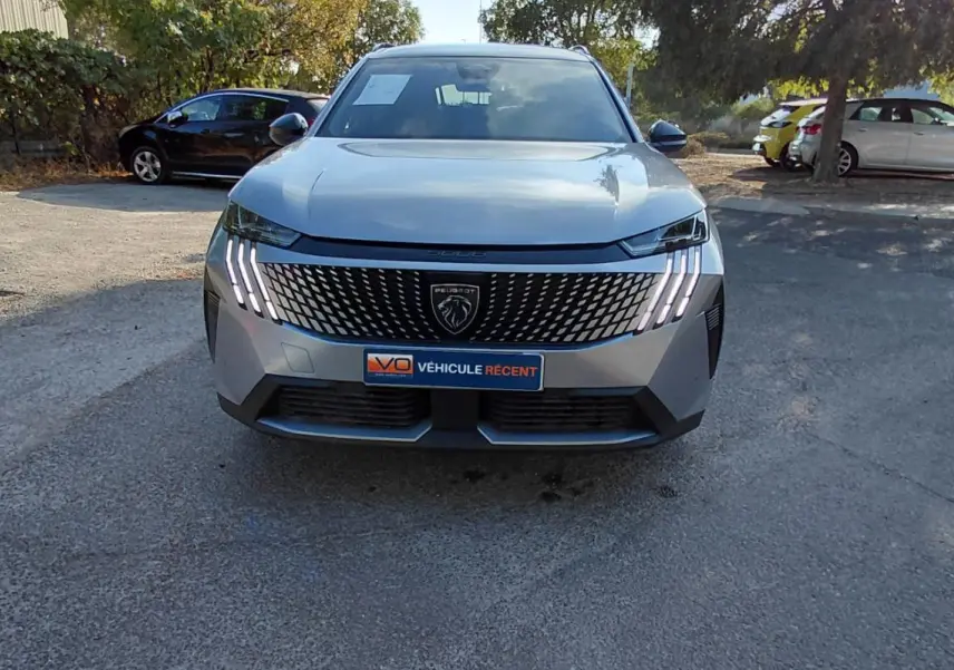 Peugeot 5008 Hybrid gris 2025 vu de face, avec calandre noire distinctive et feux LED en griffes allumés.