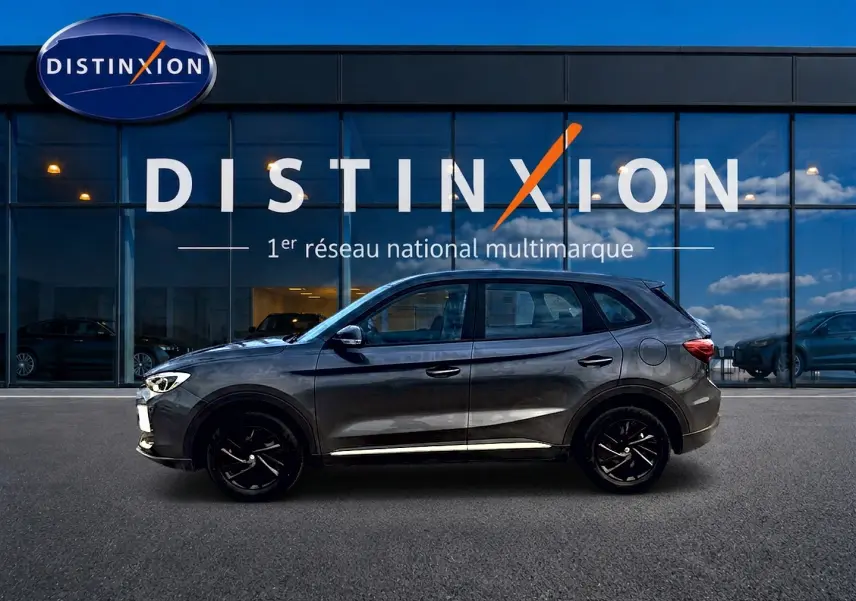 Vue de profil côté gauche du MG ZS 1.5 hybrid Camden grey devant une vitrine Distinxion, avec jantes noires et becquet arrière.