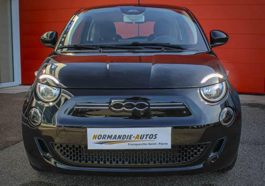 Vue frontale d'une Fiat 500 électrique noire avec logo 500 chromé sur calandre, devant un mur rouge.