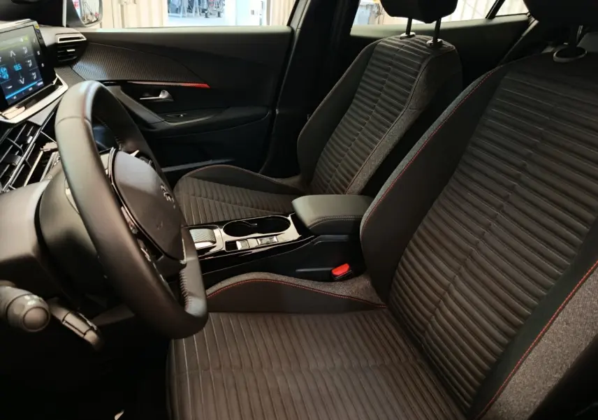 Vue intérieure côté conducteur du Peugeot E-2008 électrique, sièges noirs avec surpiqûres rouges et volant cuir multifonctions.