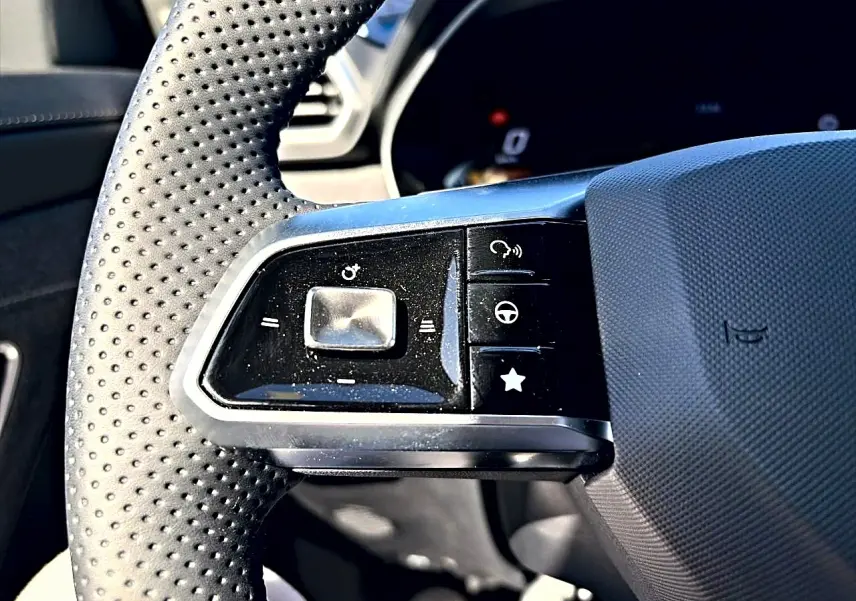 Gros plan sur les commandes gauche du volant perforé noir du MG Motor ZS 1.5 hybrid 197 standard, avec boutons multifonctions.