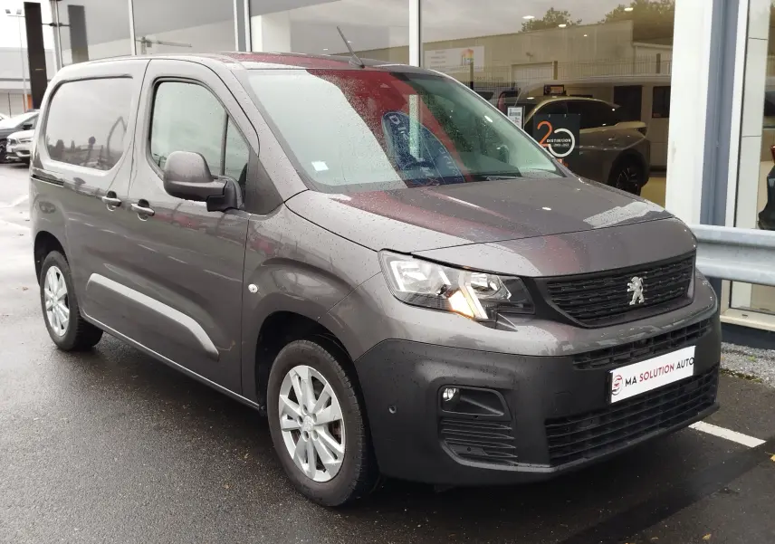 Vue 3/4 avant gauche d’un Peugeot Partner gris foncé 2020 avec phare allumé, garé devant un bâtiment vitré.