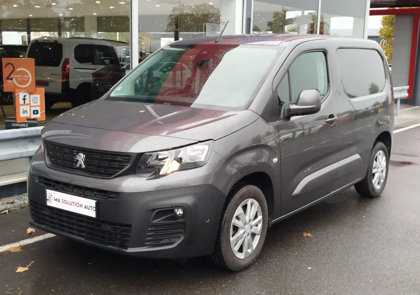 Peugeot Partner gris foncé vu en 3/4 avant droit, avec phares allumés et carrosserie utilitaire sous la pluie.