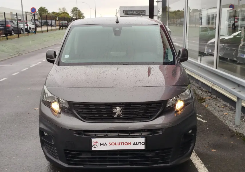 Vue de face d'un Peugeot Partner gris foncé 2020 avec feux allumés, stationné devant une vitrine sous la pluie.