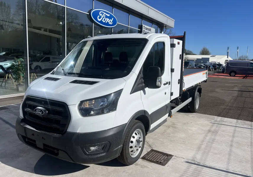 Vue 3/4 avant droit d’un Ford Transit Chassis Cabine blanc avec benne et coffre, roues jumelées, stationné devant un showroom Ford.