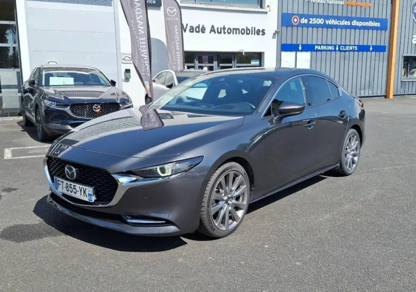 Vue 3/4 avant d'une Mazda3 gris métallisé garée devant un concessionnaire avec jantes aluminium et phares allumés.
