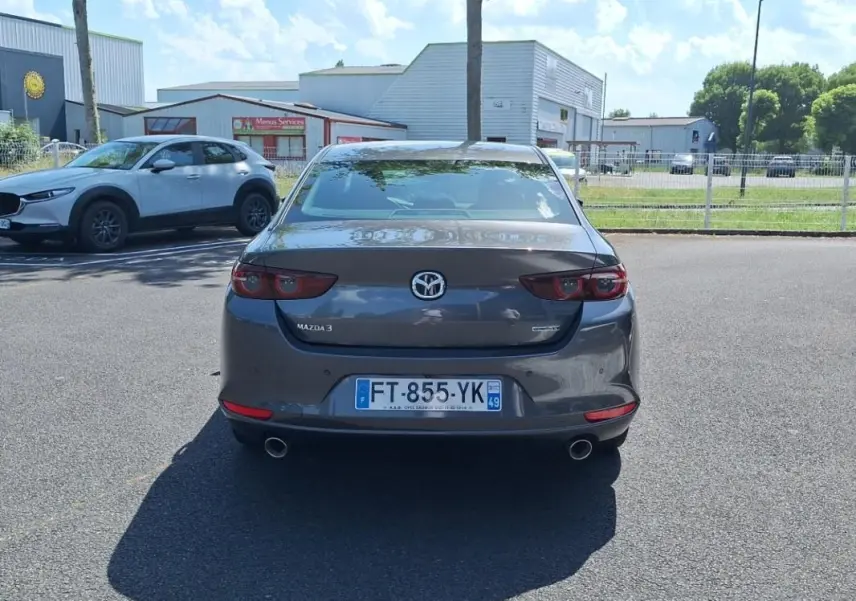 Vue arrière d'une Mazda3 gris métallisé stationnée, avec feux arrière allongés et double sortie d'échappement visible.