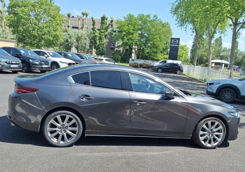 Vue de profil côté droit d'une Mazda3 gris métallisé stationnée sur un parking extérieur, avec jantes aluminium visibles.