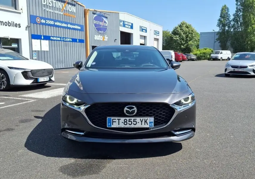 Vue avant d'une Mazda3 gris métallisé avec calandre noire et phares LED allumés, stationnée devant un bâtiment commercial.