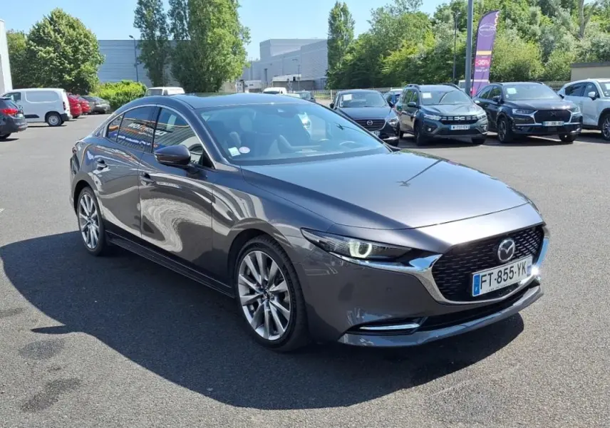 Vue 3/4 avant d'une Mazda3 gris métallisé 4 portes, avec phares allumés et jantes alliage visibles.