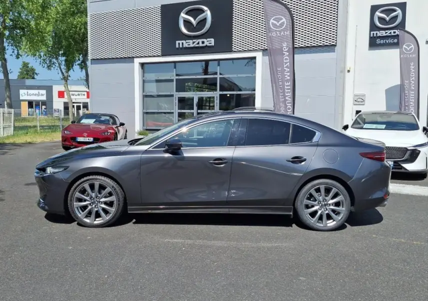 Vue latérale droite d'une Mazda3 gris métallisé garée devant une concession Mazda avec jantes alu et vitres teintées.