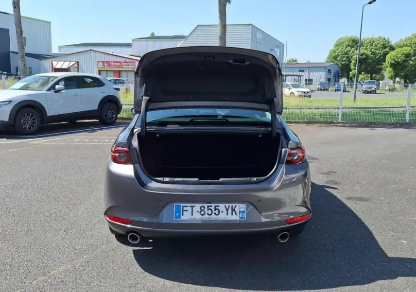 Vue arrière d'une Mazda3 gris métallisé avec coffre ouvert sur un parking extérieur ensoleillé.