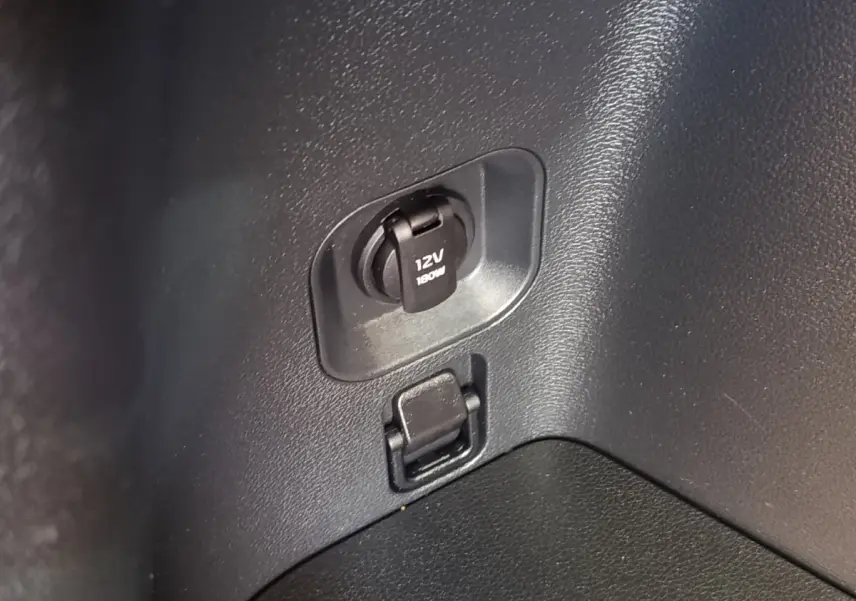 Prise 12V et port USB dans le coffre du Kia Sportage gris clair, vue rapprochée du panneau latéral intérieur.
