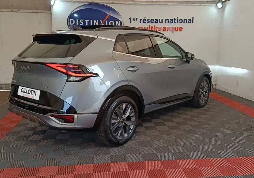 Vue 3/4 arrière droite d’un KIA Sportage gris clair avec feux arrière LED et jantes alliage foncé, en intérieur showroom.