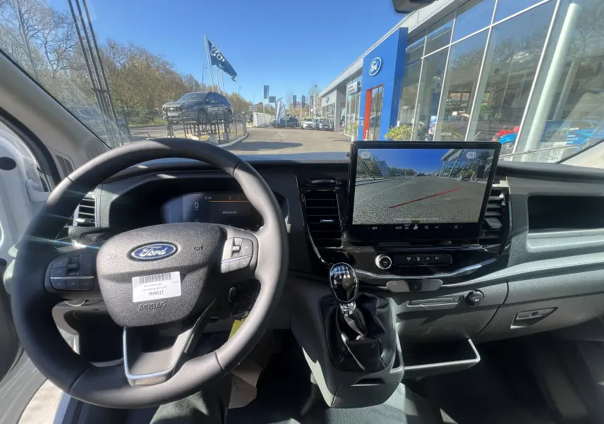 Vue intérieure du poste de conduite du Ford Transit blanc, avec volant, levier de vitesses manuel et écran central affichant la caméra de recul.