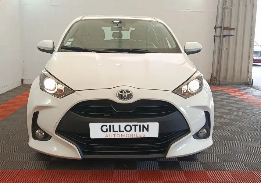 Vue frontale d'une Toyota Yaris hybride blanche avec phares allumés et calandre noire dans un showroom.
