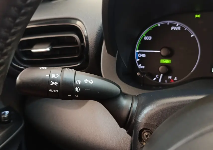 Gros plan sur la manette de commande des phares et clignotants côté gauche du tableau de bord noir de la Toyota Yaris hybride blanche.